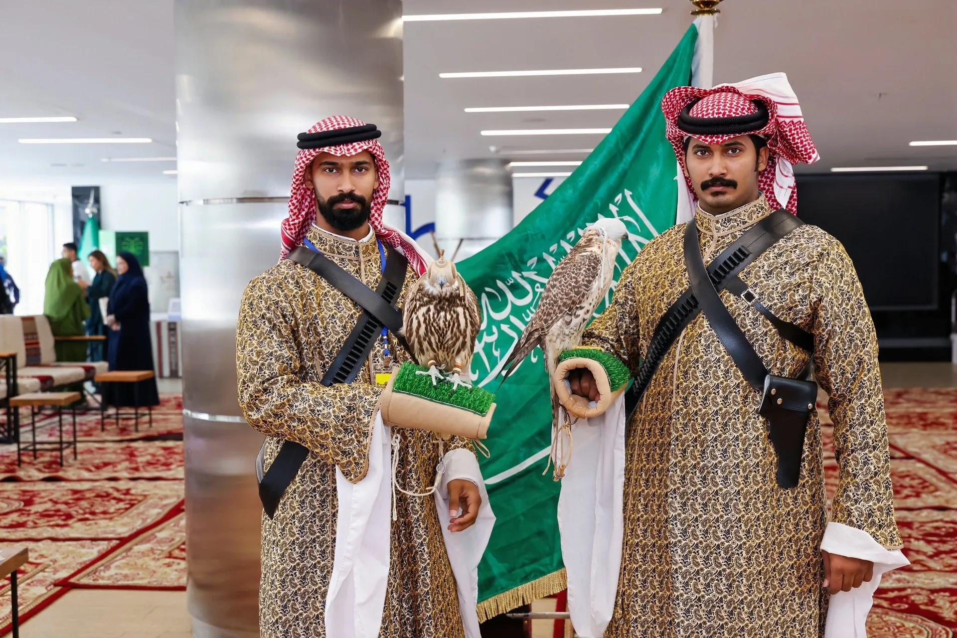 Two costumed falconers with falcons before Saudi flag