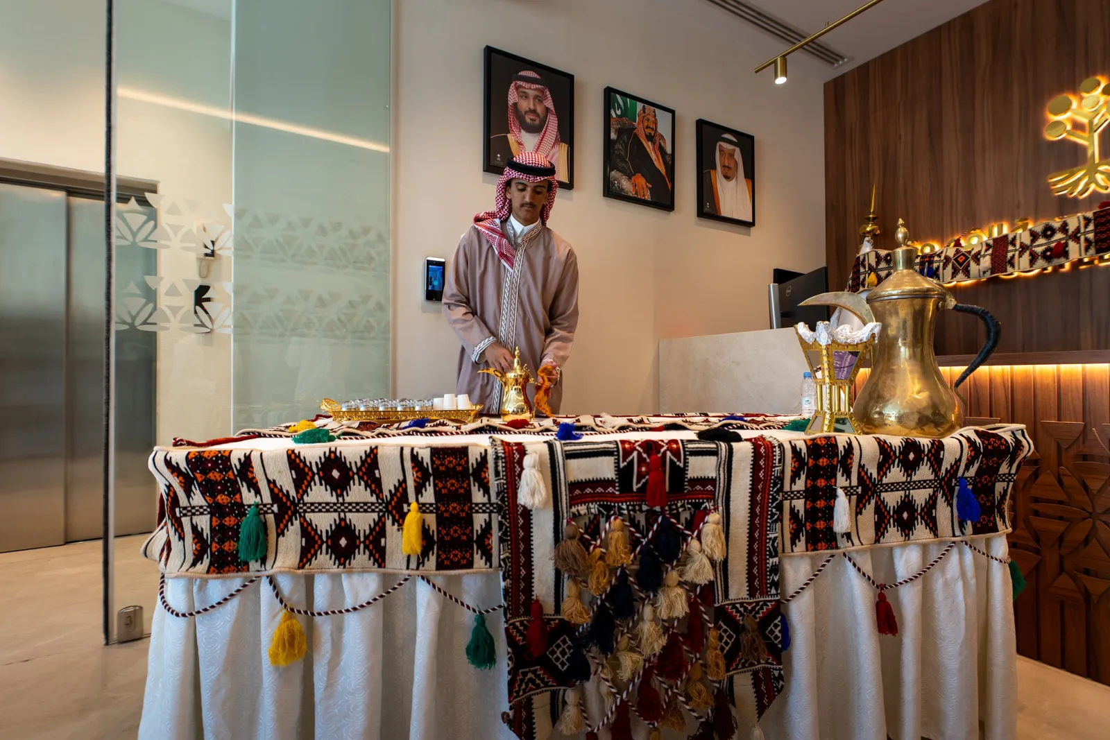 Host pouring Saudi coffee at sadu-draped table under leaders portraits